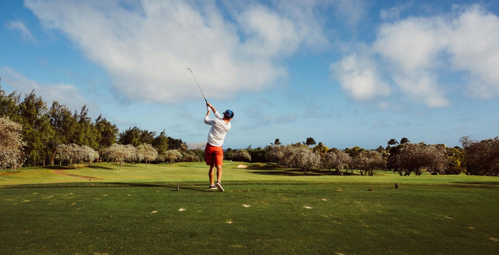 Man swinging golf club on golf course wearing white shirt and red shorts